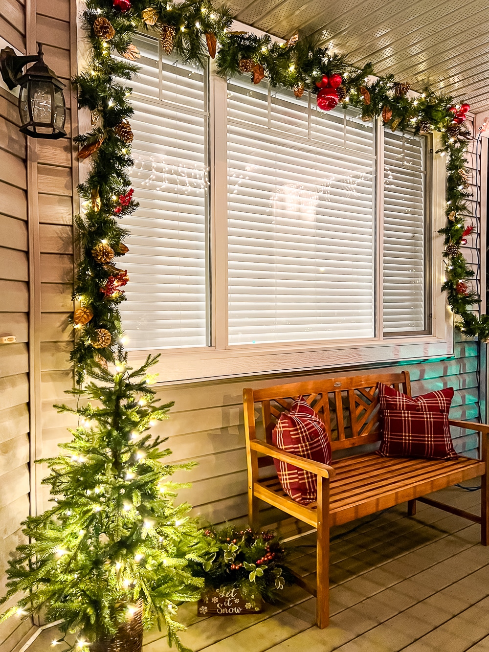 A porch festively decorated with garland around the window and a mini Chrsitmas tree with twinkle lights.